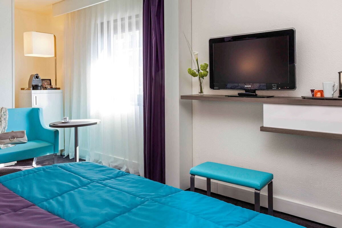 Habitación de hotel con una cama, zona de estar con una silla y una mesita, televisor sobre un mueble y una ventana con cortinas.