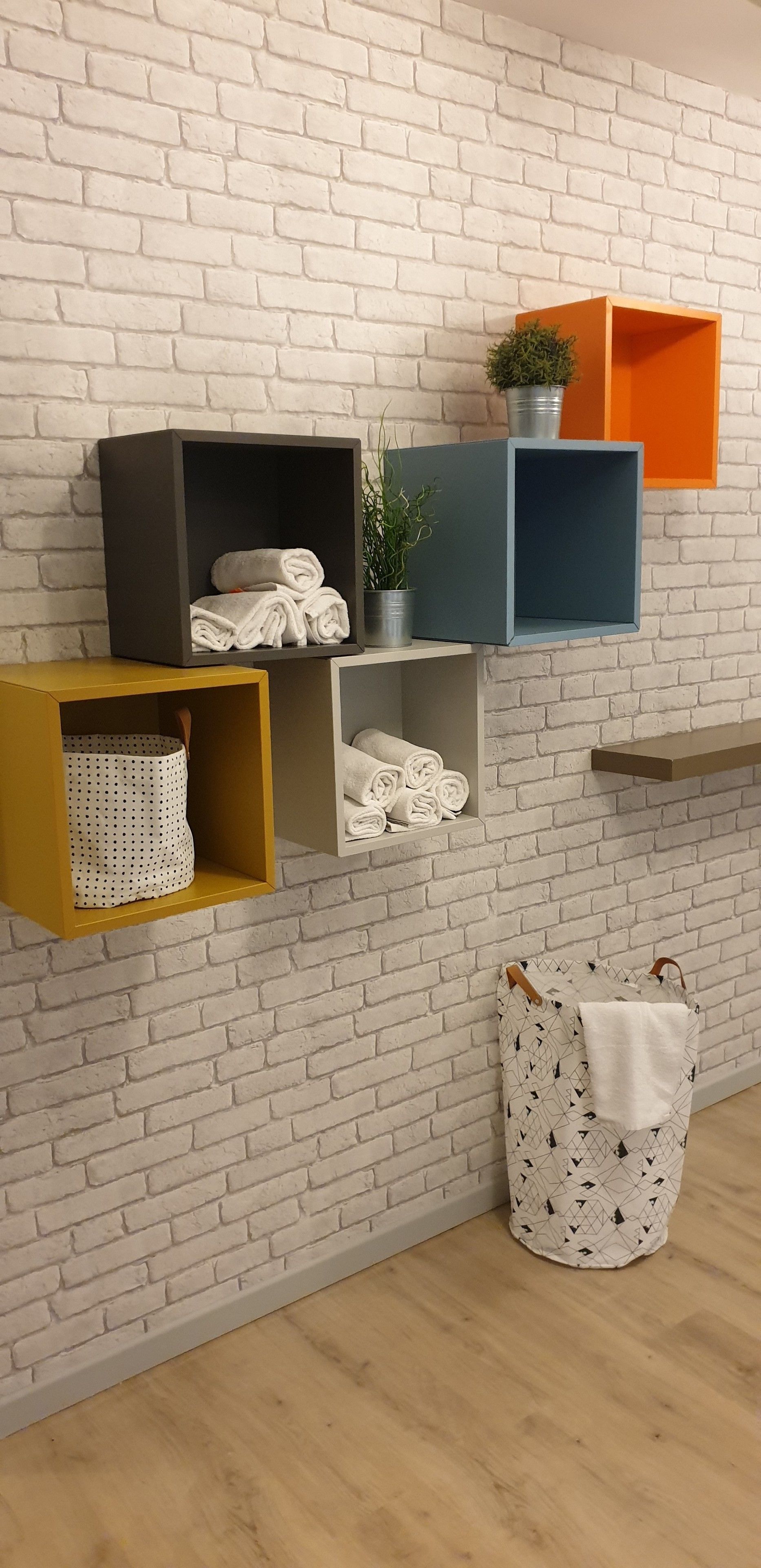 A room with a white brick wall, shelves with folded towels, a potted plant, and a hanging bag.