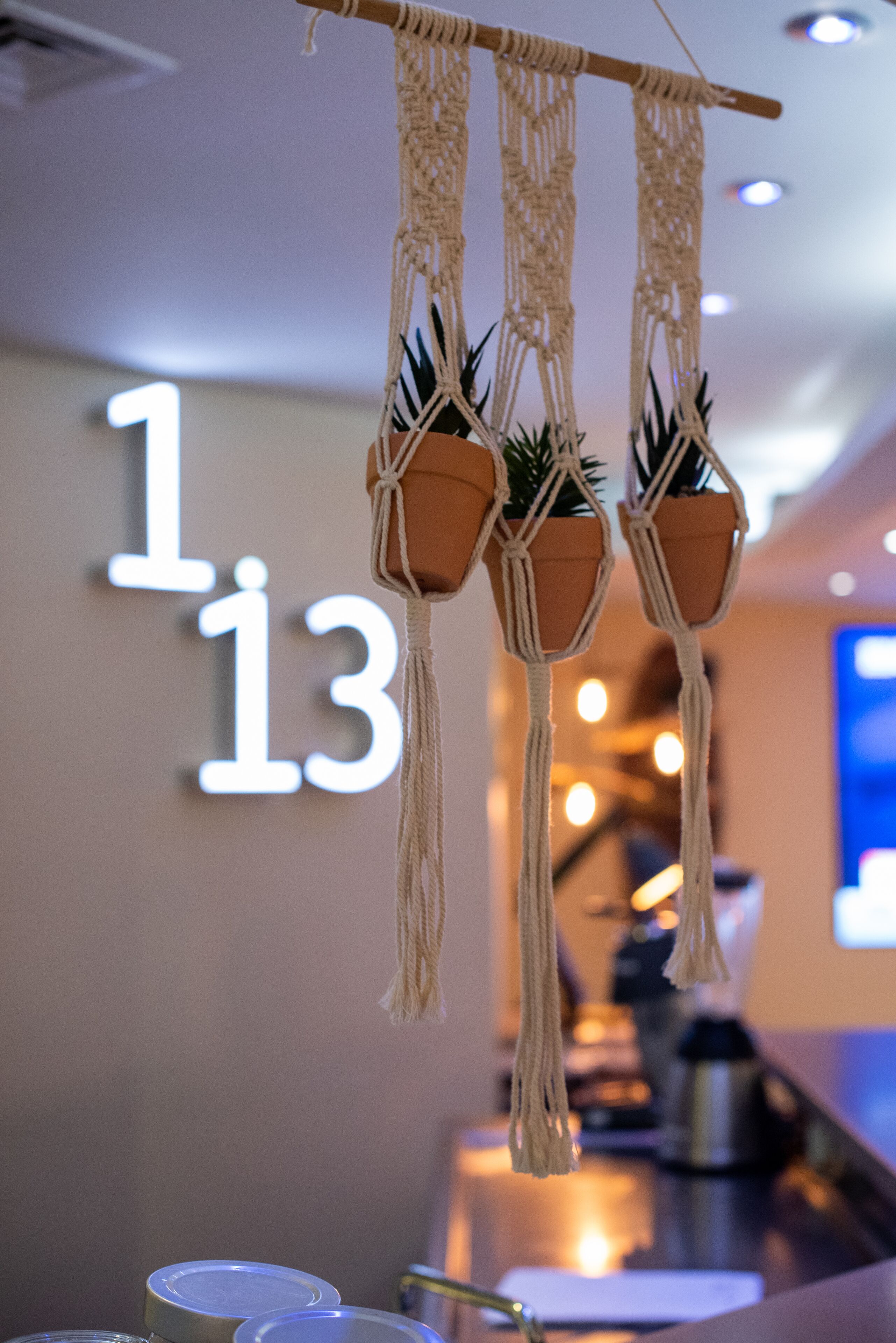 A macrame wall hanging with three terracotta pots containing plants, a number '13' illuminated sign in the background, and a bar area with a coffee machine.