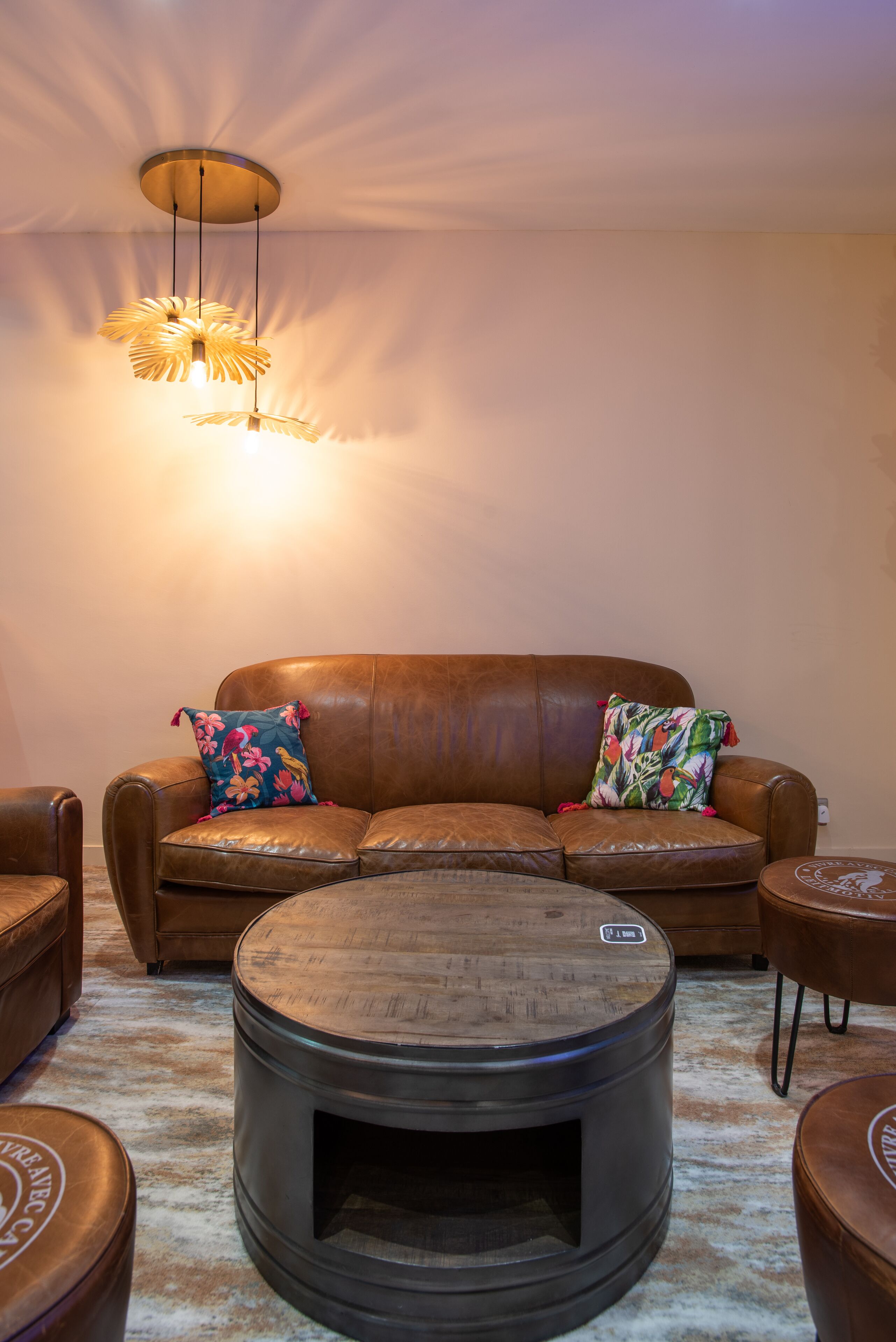 A living room with a brown leather sofa, two colorful pillows, a round wooden coffee table, and a hanging light fixture.