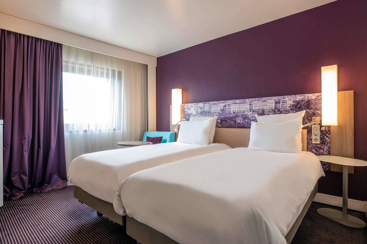 A hotel room with two beds, a small table, a chair, and a window with curtains.