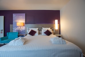 A hotel room with a large bed, two pillows, a headboard, a nightstand, a lamp, a chair, and a painting on the wall.