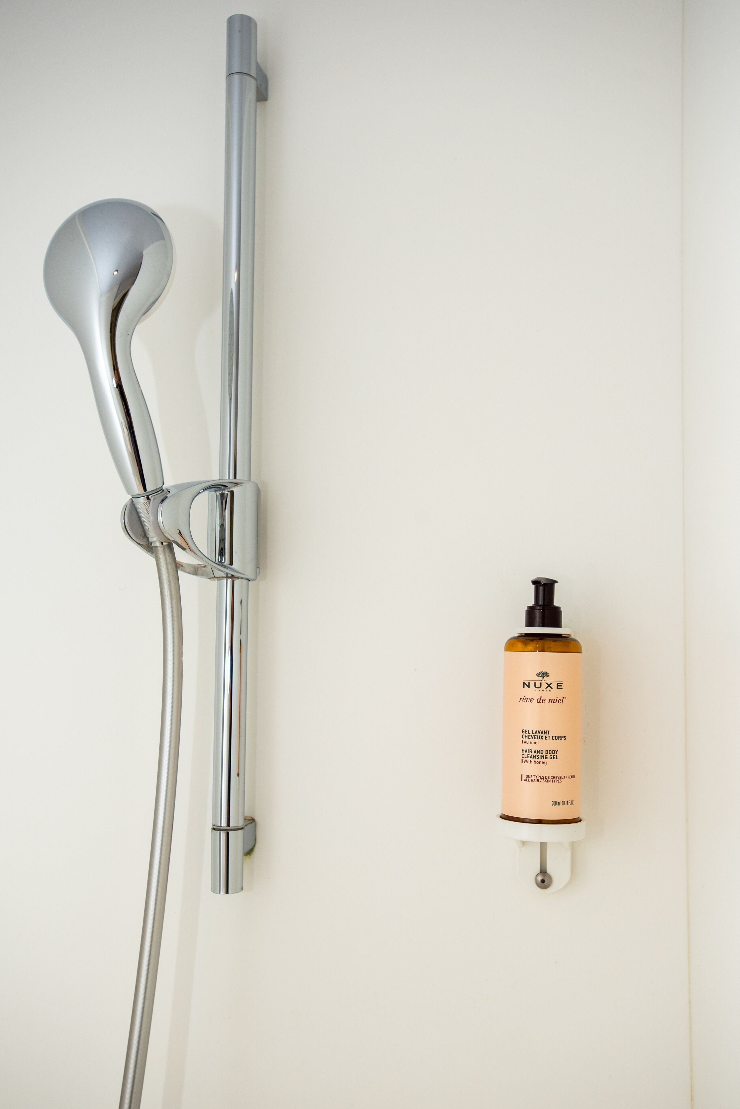 Ducha con grifo de cromo y una botella de gel limpiador corporal Nuxe colgada en la pared.