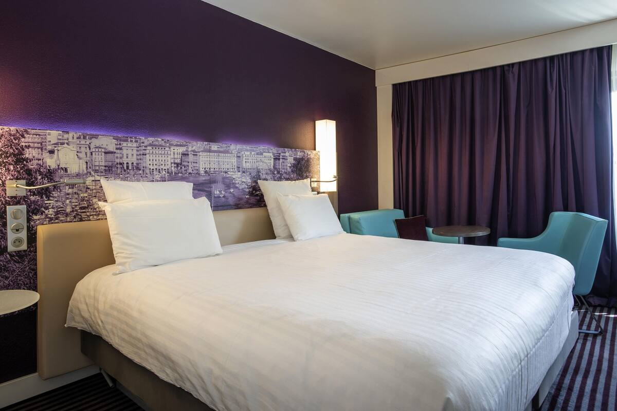 A hotel room with a large bed, two chairs, a small table, a cityscape mural, and a window with curtains.