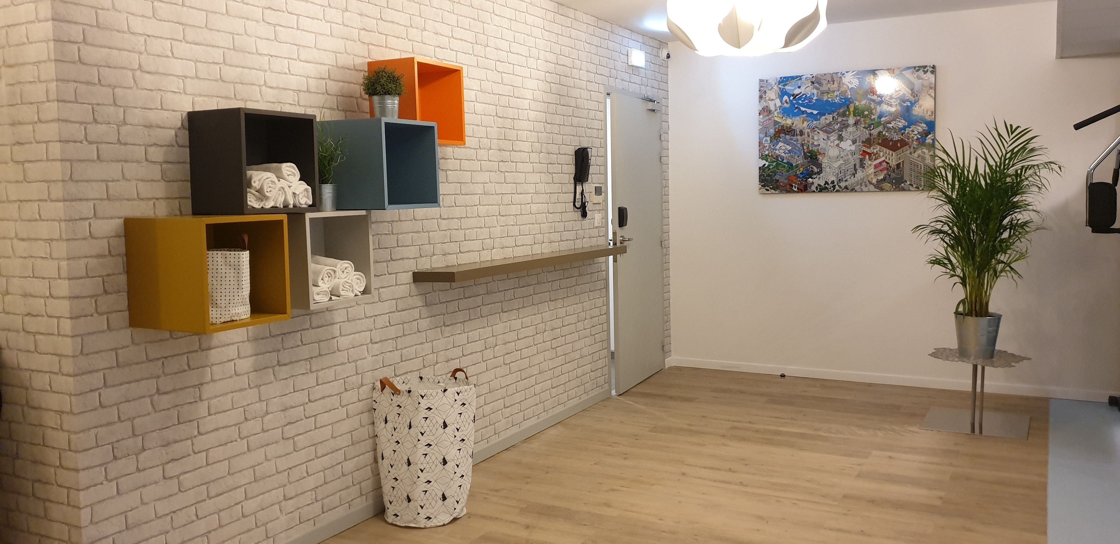 A room with a white brick wall, wooden flooring, and a door. There are shelves with various items, a potted plant, and a painting on the wall.