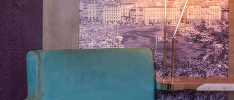 Un fauteuil de couleur turquoise dans une pièce ornée d’une fresque représentant un paysage urbain, avec un lampadaire et un panneau en bois sur le mur.
