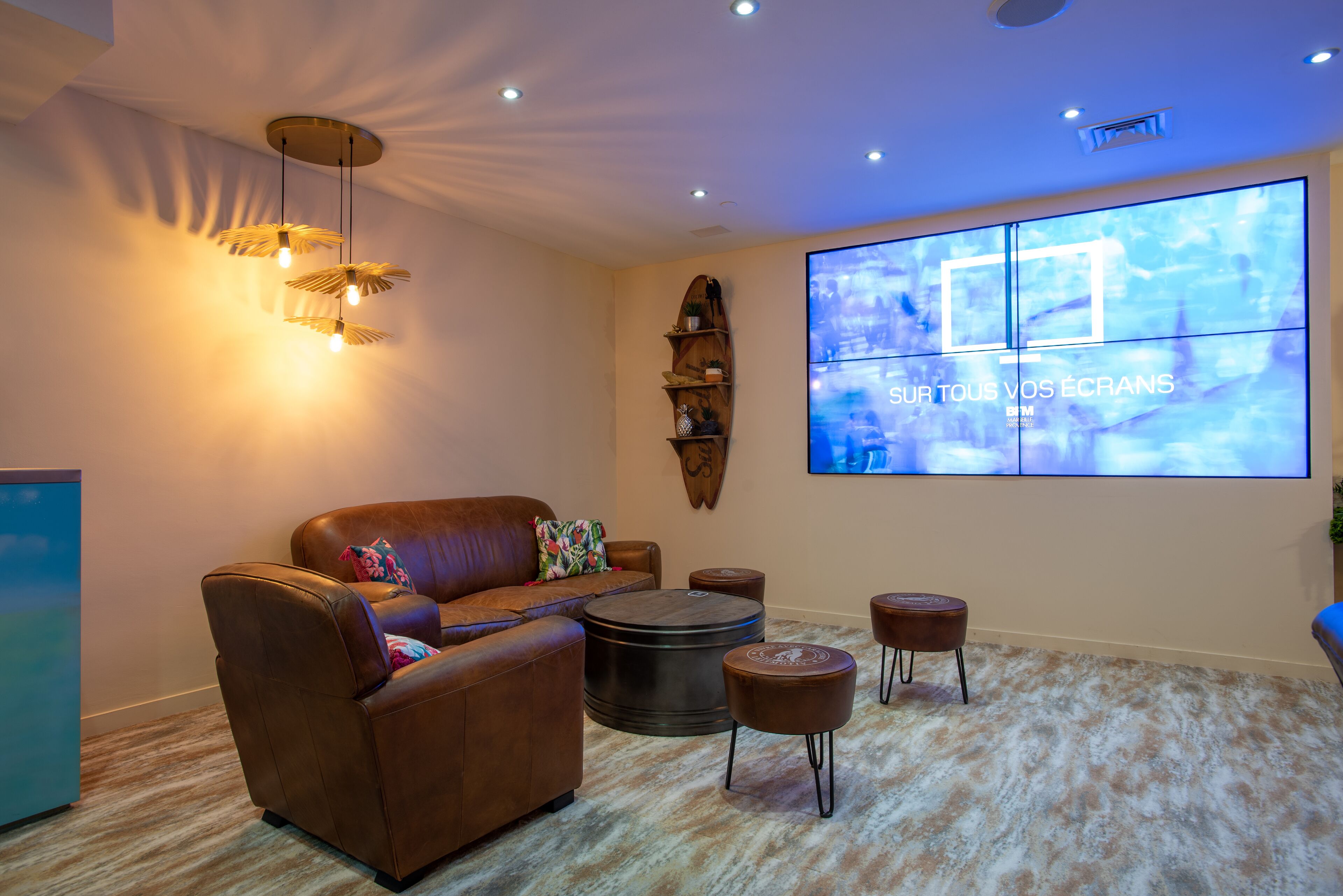 Un salon moderne avec un canapé en cuir, une table basse ronde et un pouf, un grand téléviseur à écran plat et un luminaire suspendu au plafond.