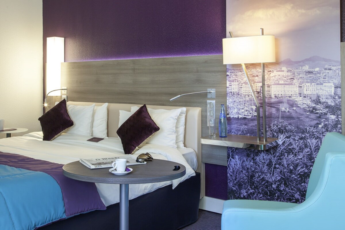 Habitación de hotel con cama, una mesa redonda con taza y platillo, una silla y vista a la ciudad.
