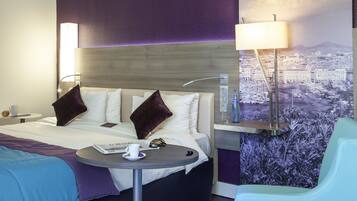 A hotel room with a bed, a round table with a cup and saucer, a chair, and a cityscape view.