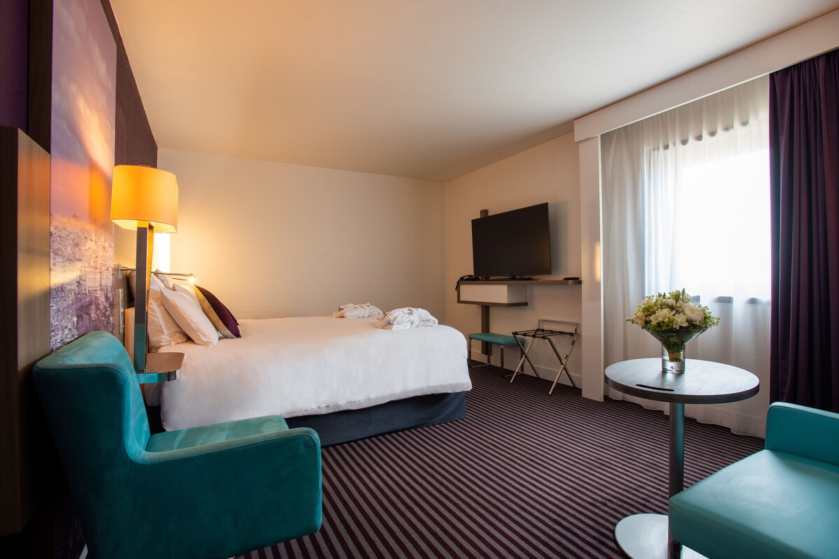 Habitación de hotel con cama, escritorio, silla, televisor y una pequeña mesa con un florero.