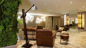 A modern living room with a leather sofa, a coffee table, and a floor lamp. There is a large green wall with plants and a glass partition with a decorative display.