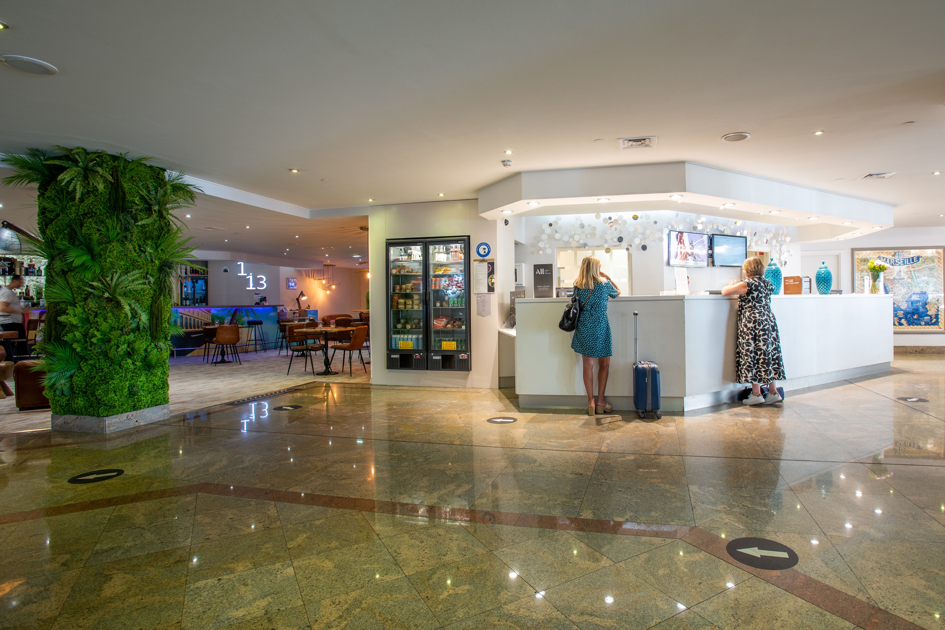 Un vestíbulo de hotel con un mostrador de recepción, una planta verde grande y una zona de bar al fondo.