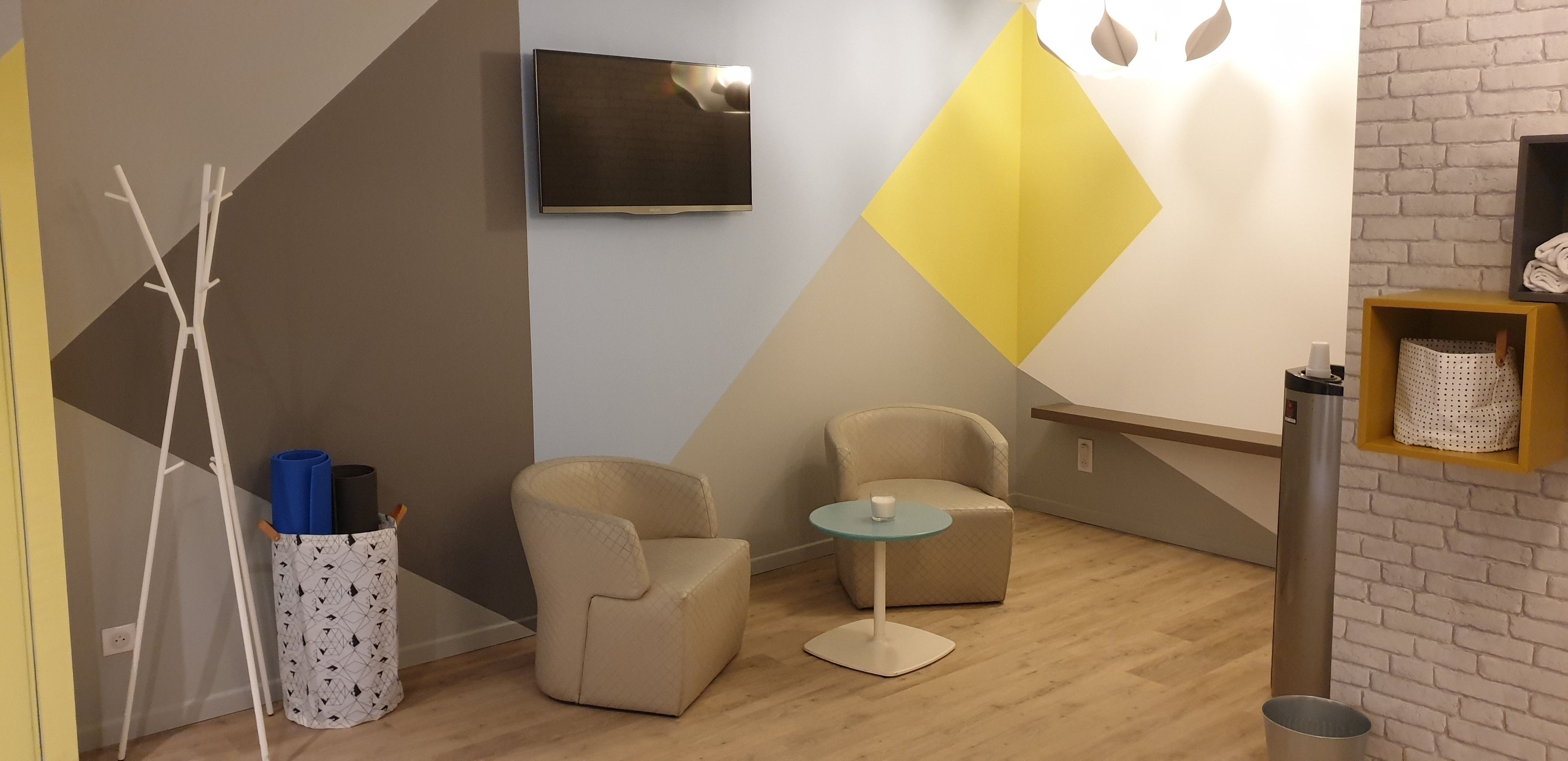 A modern room with two beige armchairs, a small round table, a TV mounted on the wall, and a brick wall with a shelf.
