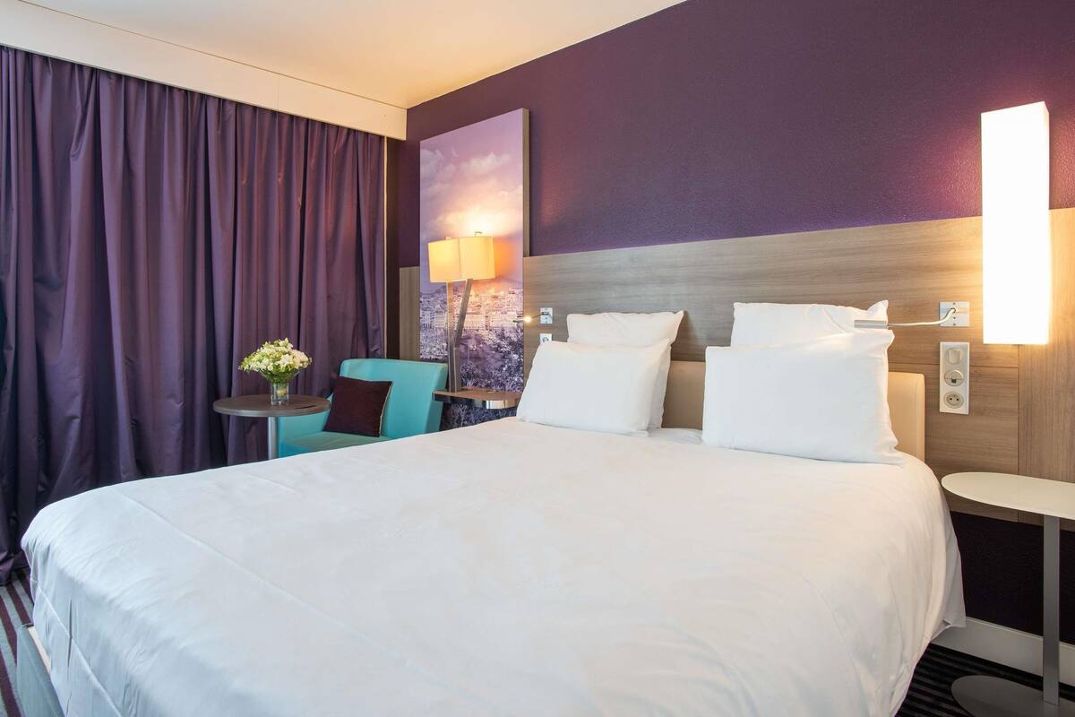 A hotel room with a large bed, a desk with a chair, a lamp, and a view of a cityscape.