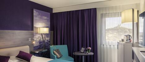 Habitación de hotel con cama, una silla, una mesita, un escritorio y una ventana con cortinas.