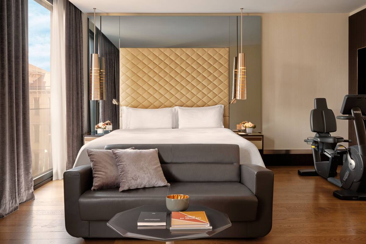 A modern hotel room with a large bed, a sofa, a treadmill, and a coffee table with books.