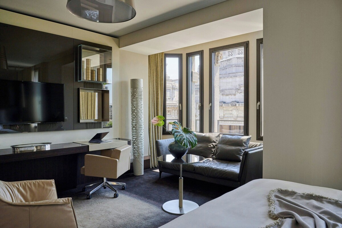 A modern hotel room with a large bed, a desk with a computer, a sofa, and a large window offering a city view.