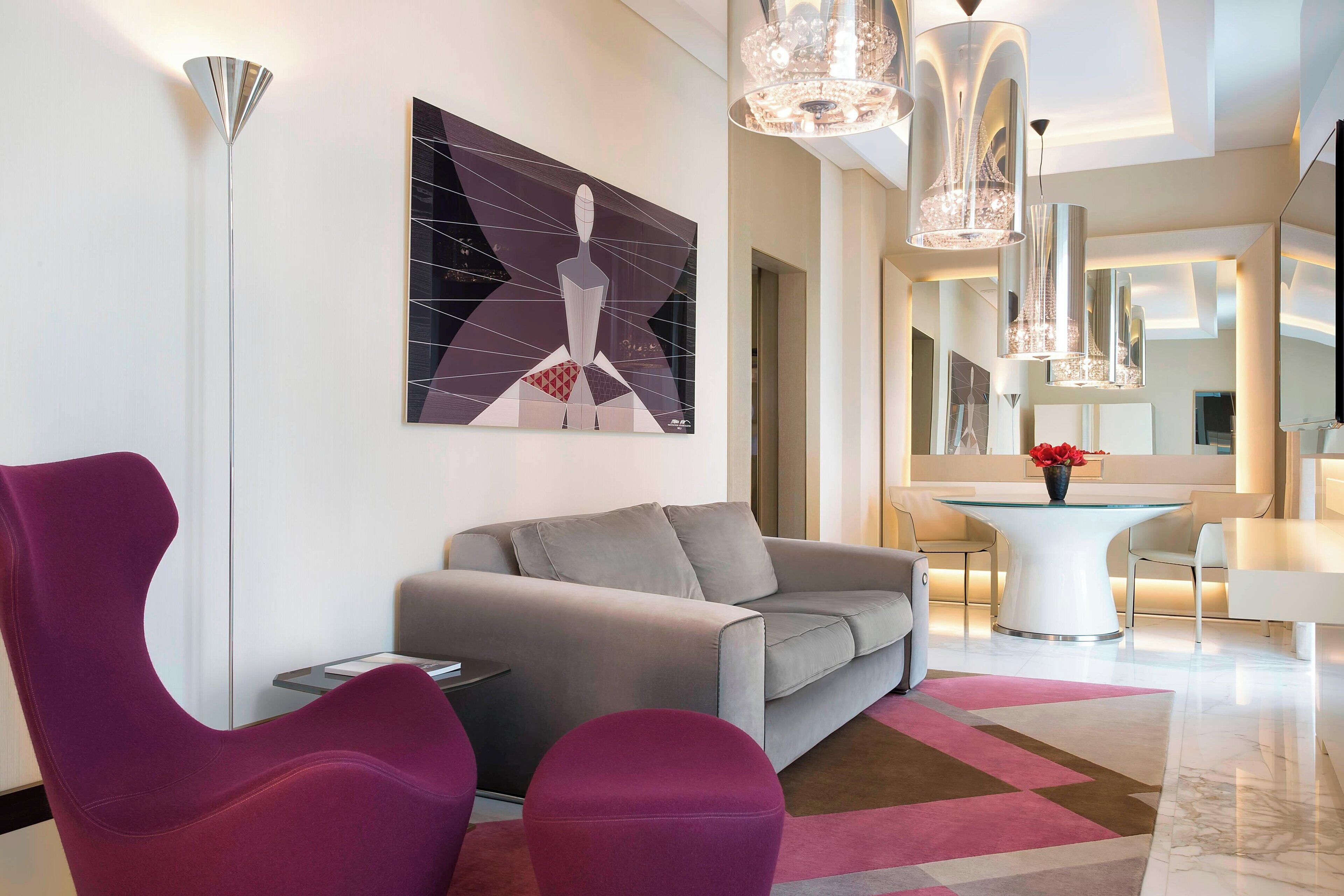 Een moderne woonkamer met een grijze bank, paarse fauteuils, een glazen salontafel en een groot abstract schilderij.