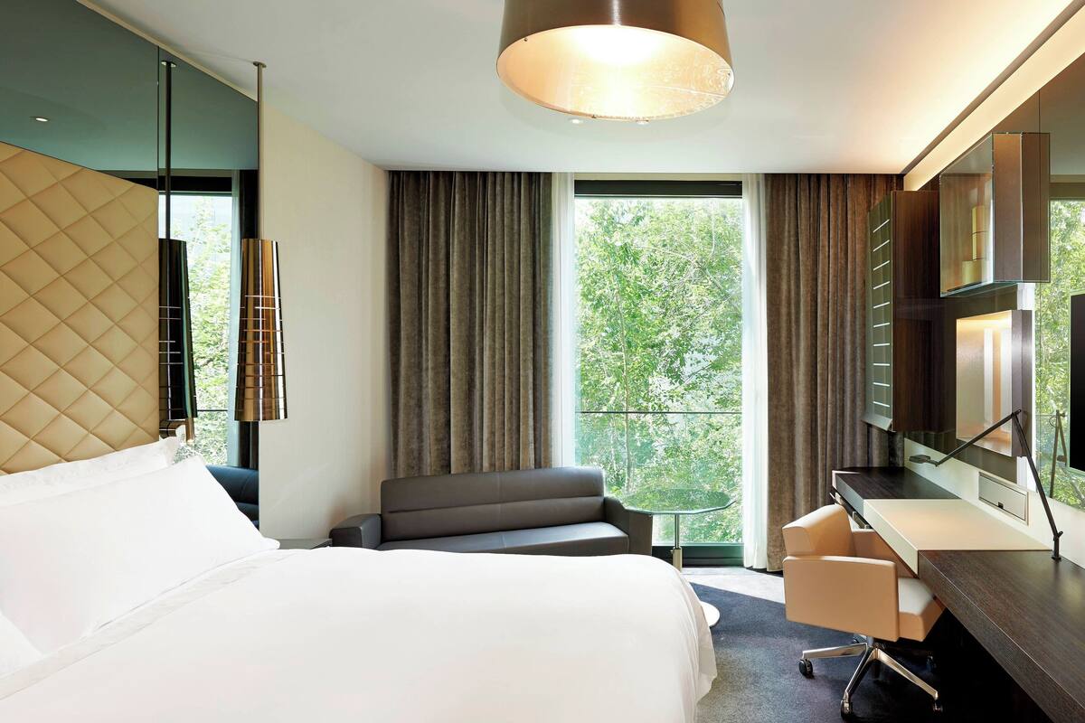 A hotel room with a bed, a sofa, a desk with a chair, a TV, and a large window with curtains.