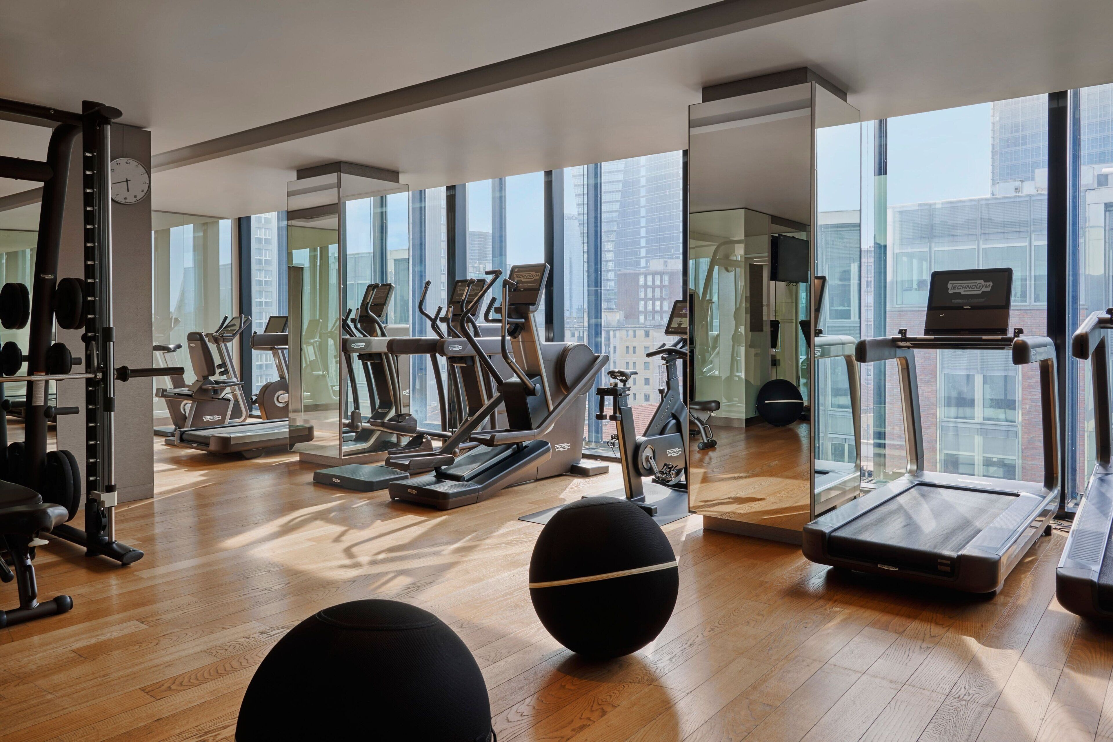 Una palestra moderna con diverse attrezzature per l'esercizio fisico, ampie finestre e pavimento in legno.