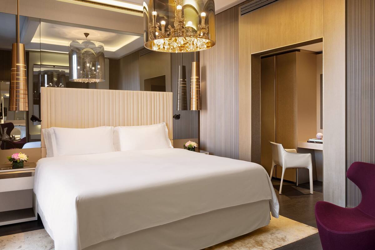 A modern hotel room with a large bed, a desk, a chair, and a chandelier.