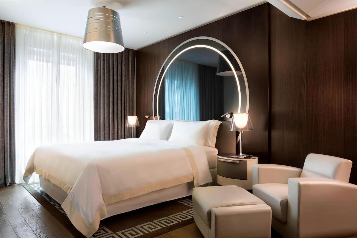 A modern hotel room with a large bed, bedside lamps, a round mirror, and a seating area with a chair and ottoman.