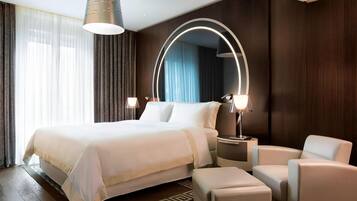 A modern hotel room with a large bed, bedside lamps, a round mirror, and a seating area with a chair and ottoman.