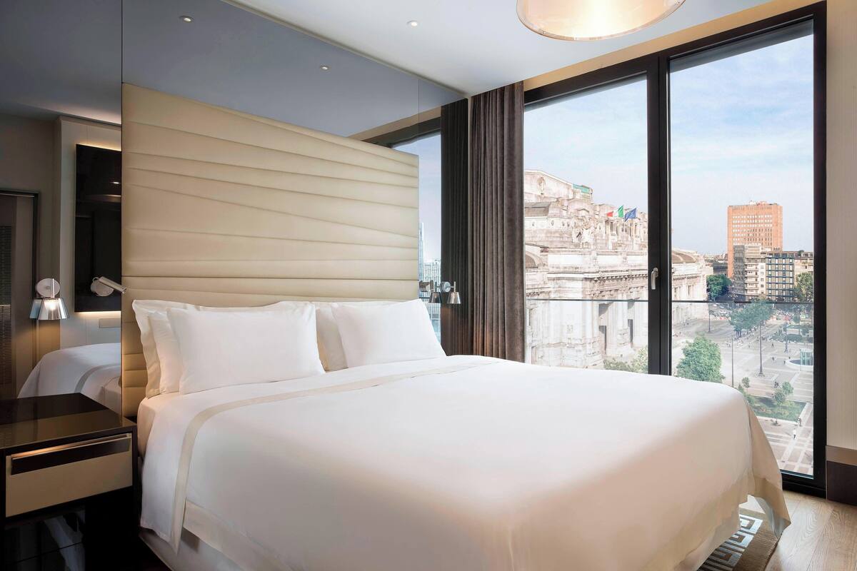 A modern hotel room with a large bed, a nightstand, and a view of a cityscape through large windows.
