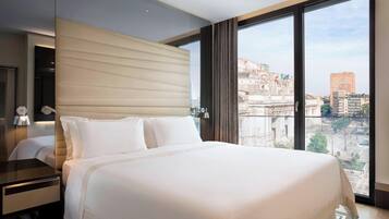 Ein modernes Hotelzimmer mit einem großen Bett, einem Nachttisch und Blick auf eine Stadtlandschaft durch große Fenster.