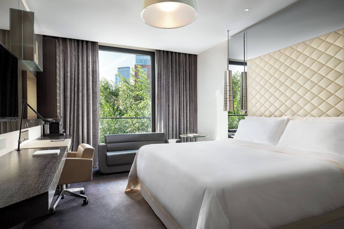 A modern hotel room with a large bed, a desk with a chair, a sofa, and a view of greenery outside.