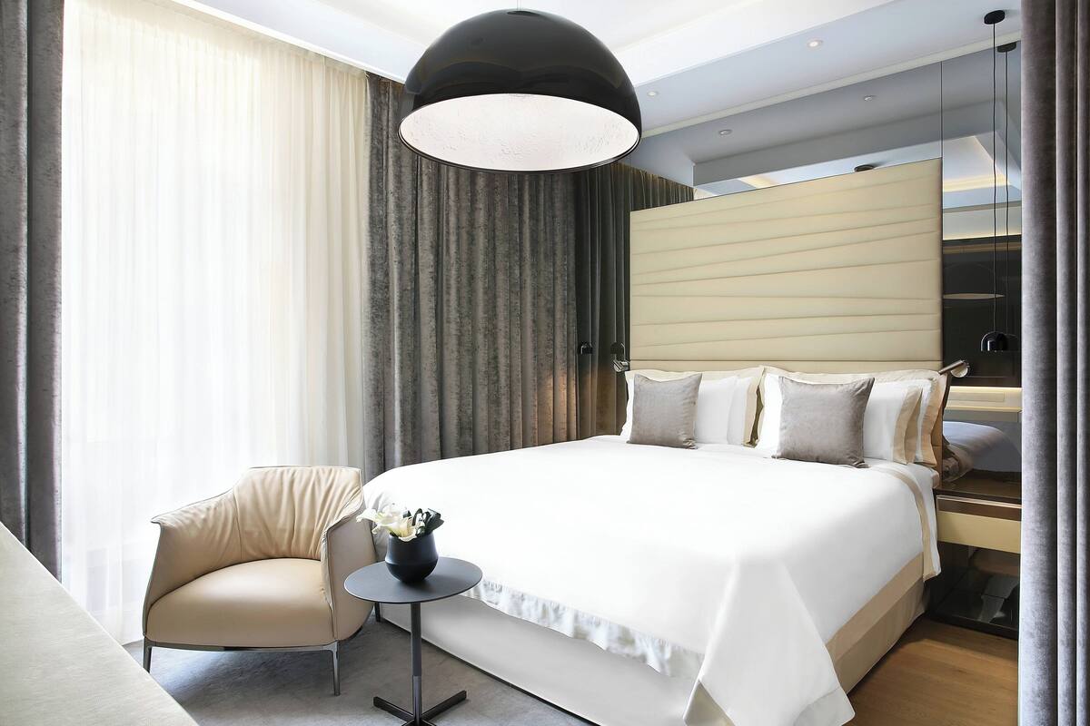 A modern hotel room with a large bed, a small round table, a chair, and a large window with curtains.