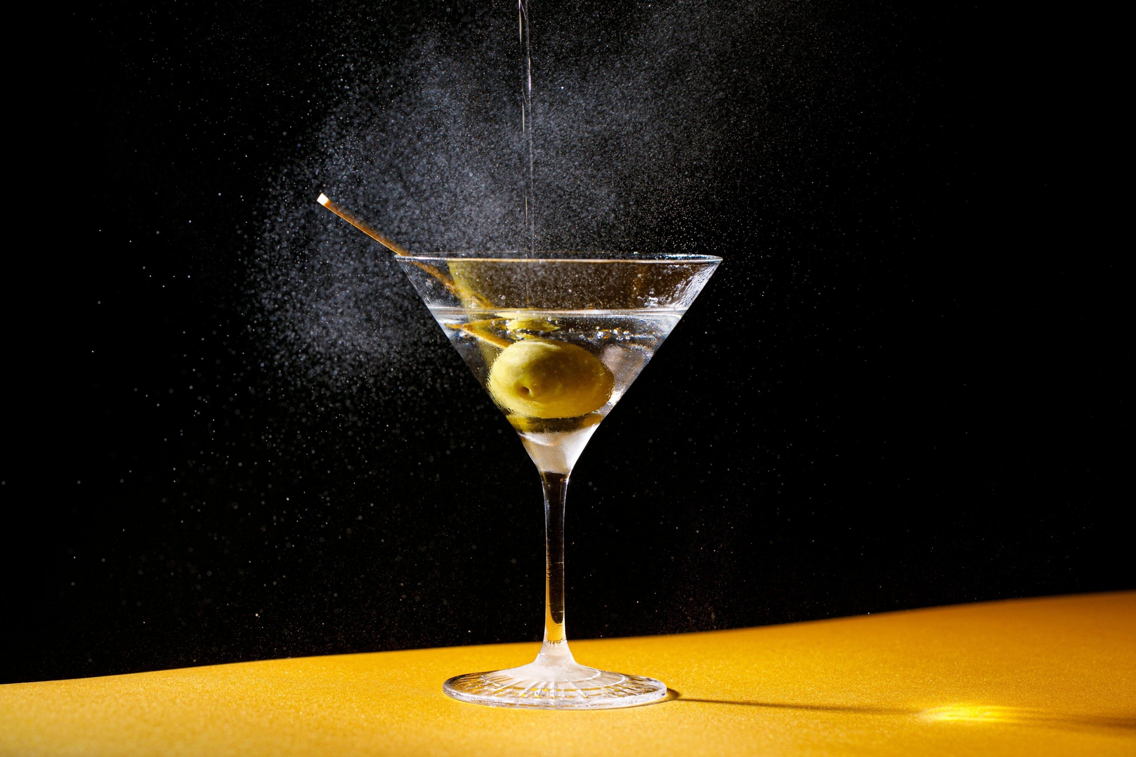 A martini glass with a lemon slice and a straw, set against a dark background with a yellow surface below.
