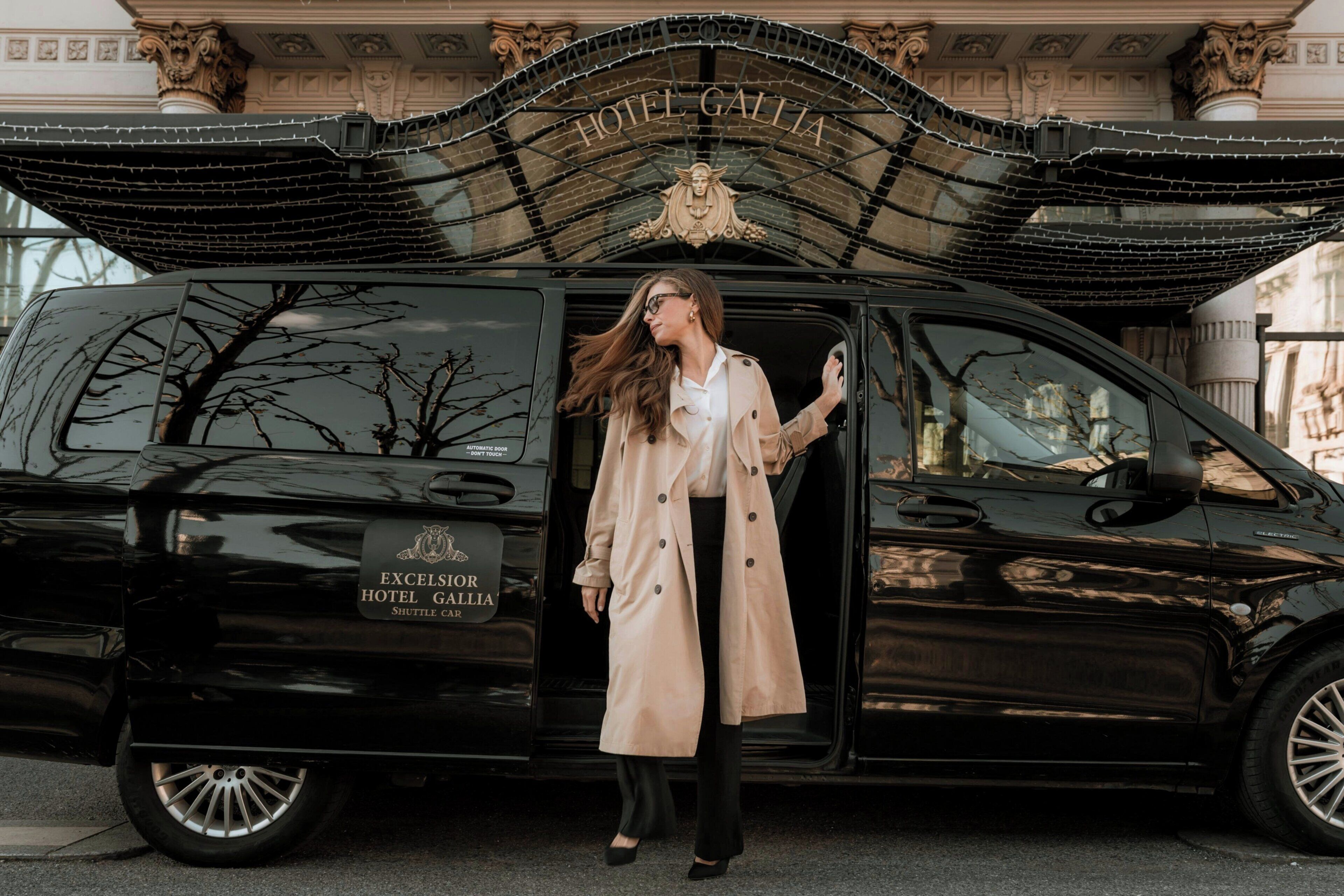 Une navette électrique noire, portant l’inscription « Excelsior Hotel Gallia », est garée devant un bâtiment à l’architecture classique.