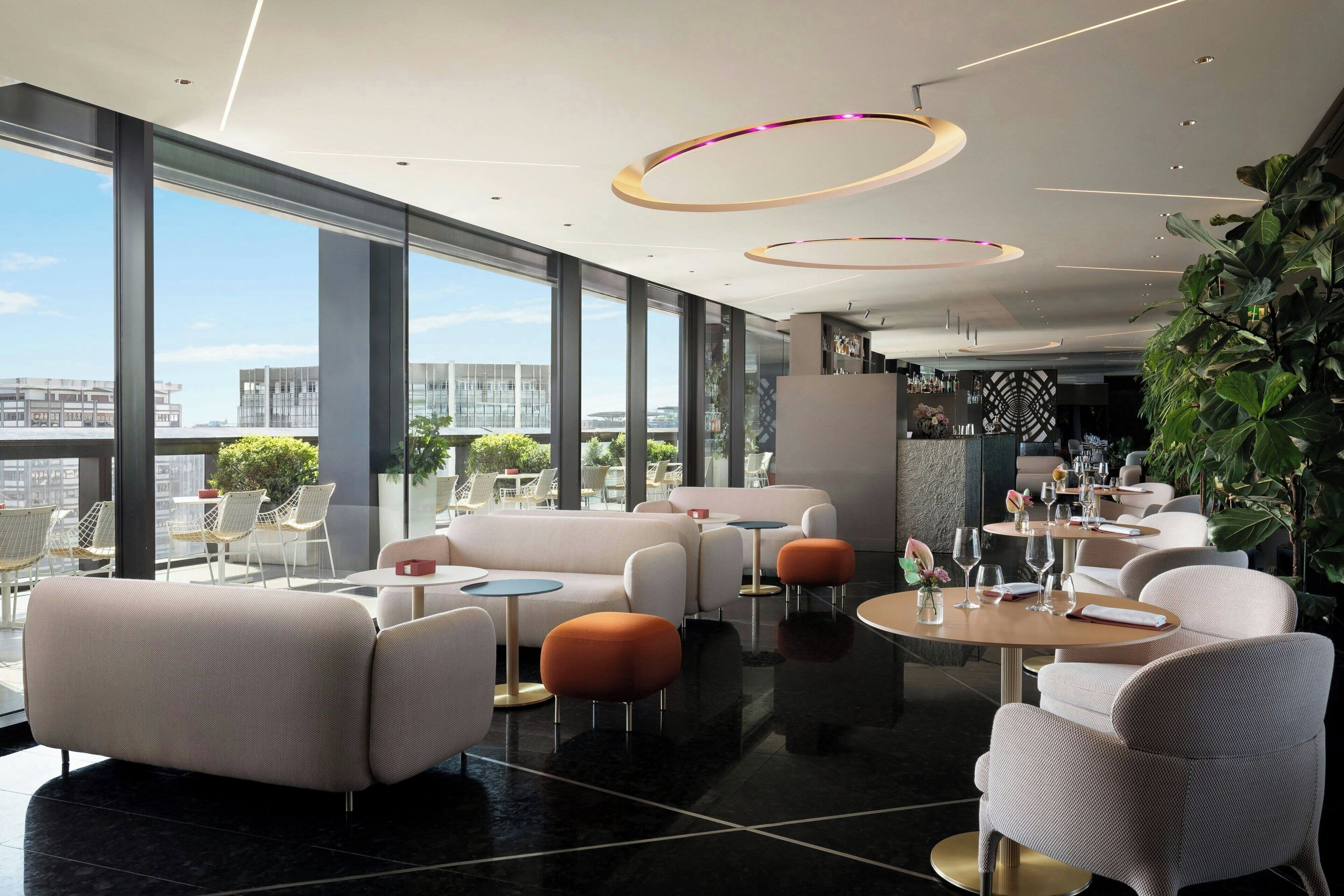 Una sala lounge moderna con divani, tavoli rotondi e ampie finestre che offrono vista sulla città.