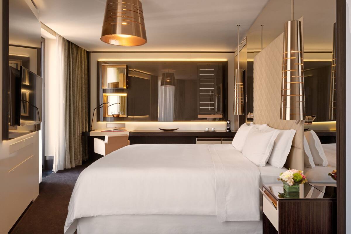 A hotel room with a large bed, a desk with a mirror, a TV, and a modern chandelier.