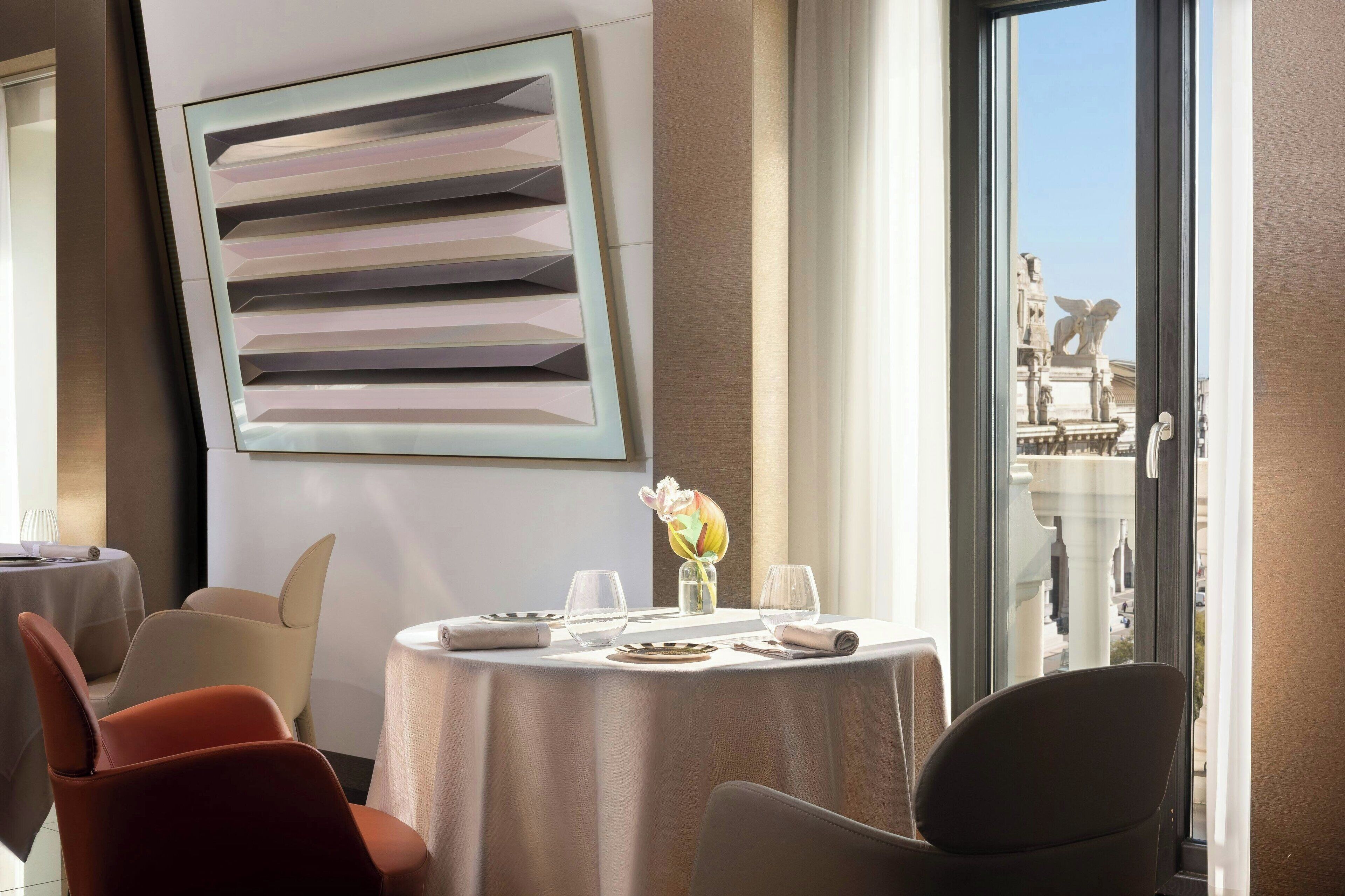 Un coin repas avec une table dressée pour deux personnes, un vase rempli de fleurs et une vue sur un bâtiment par la fenêtre.
