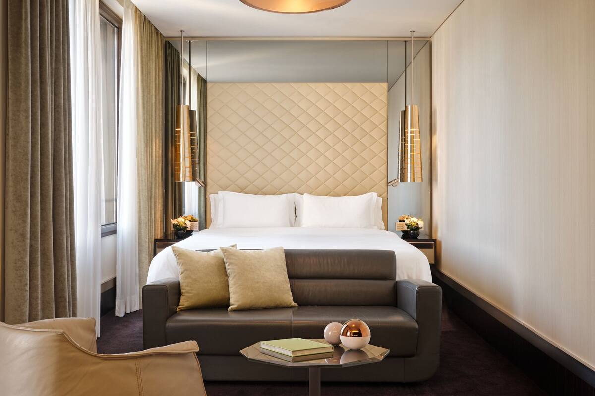 A modern hotel room with a large bed, a sofa, a small table with a book and a decorative item, and a mirror on the wall.