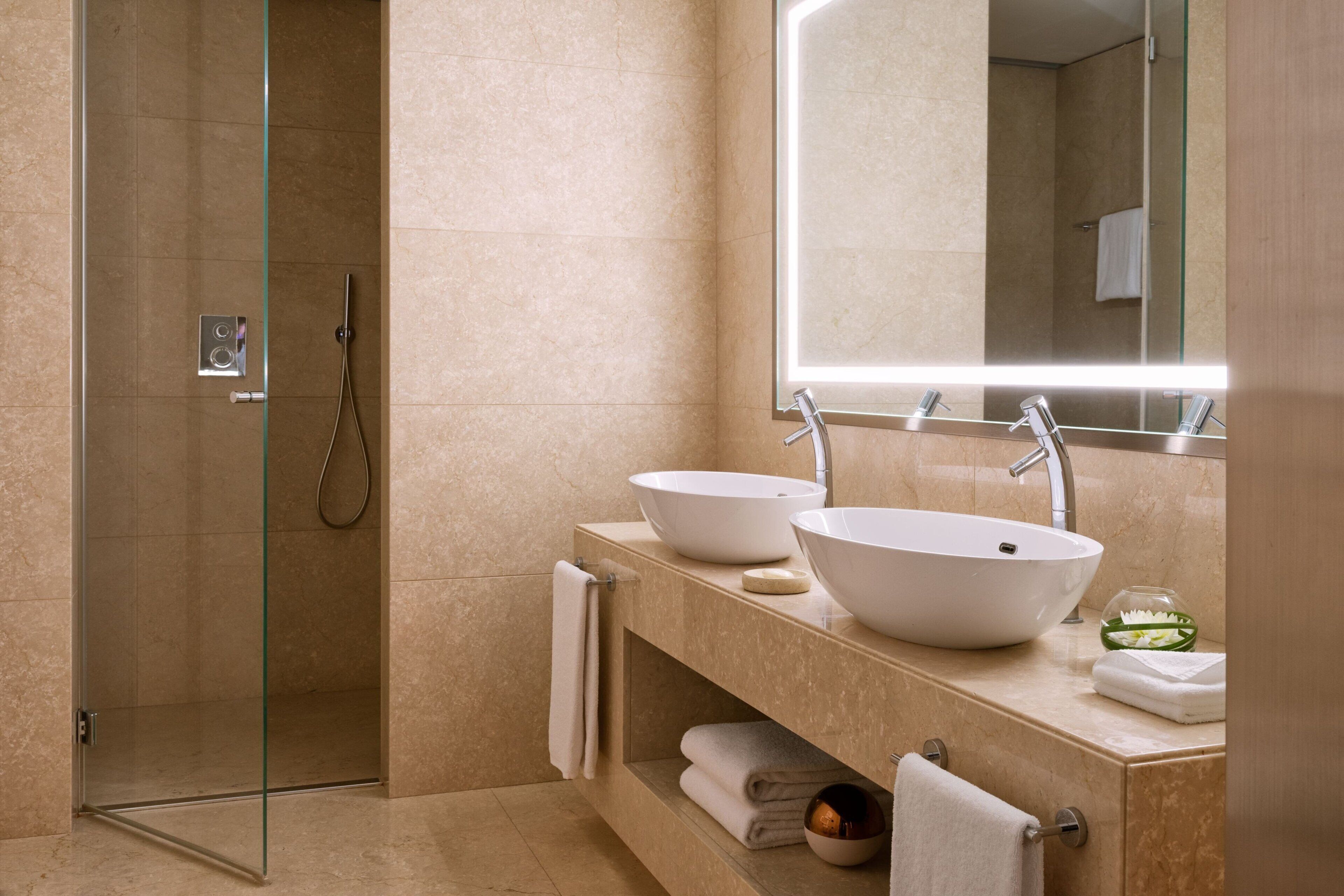 Een moderne badkamer met een douche in een glazen cabine, twee wasbakken op zuil, een grote spiegel en een marmeren blad.
