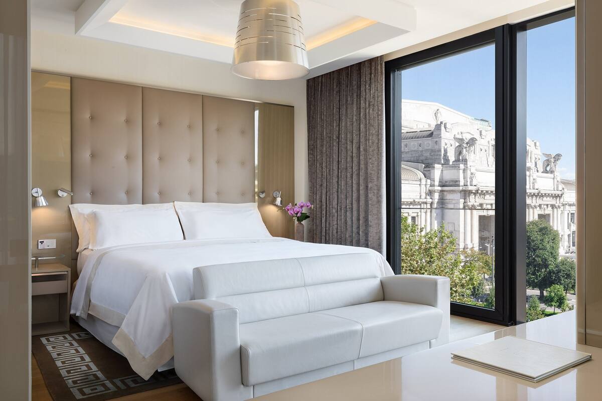 A modern hotel room with a large bed, a sofa, a nightstand, and a view of a historic building.