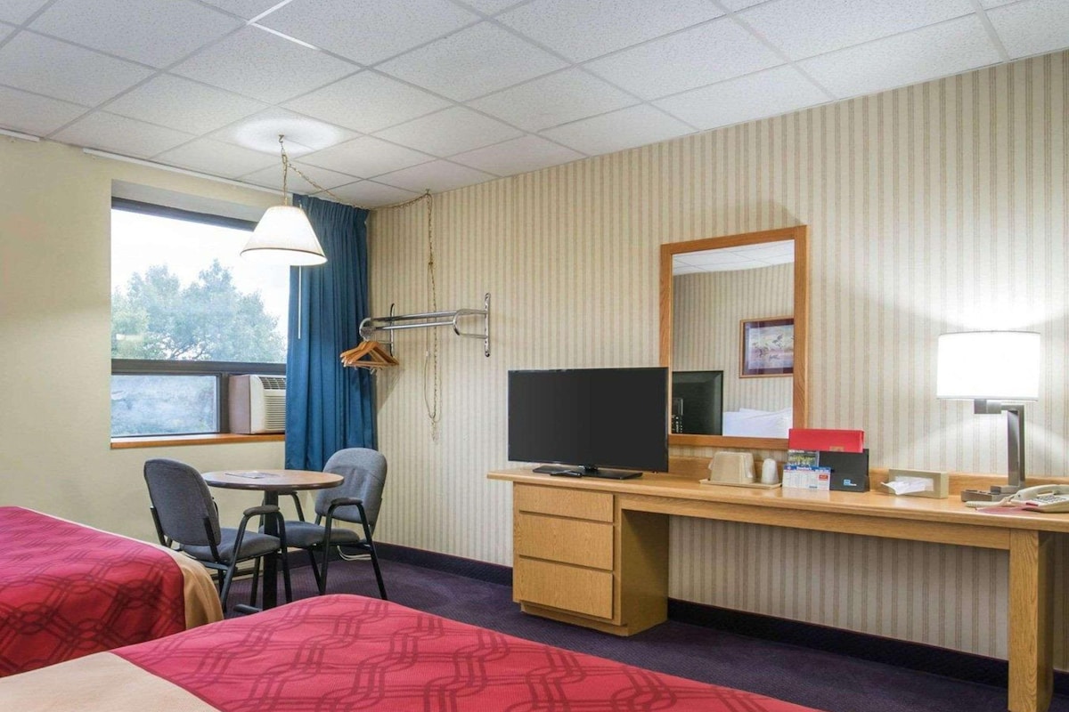 Ein Hotelzimmer mit zwei Betten, einem Schreibtisch mit Computer, einem Fernseher und einem Fenster mit Vorhängen.