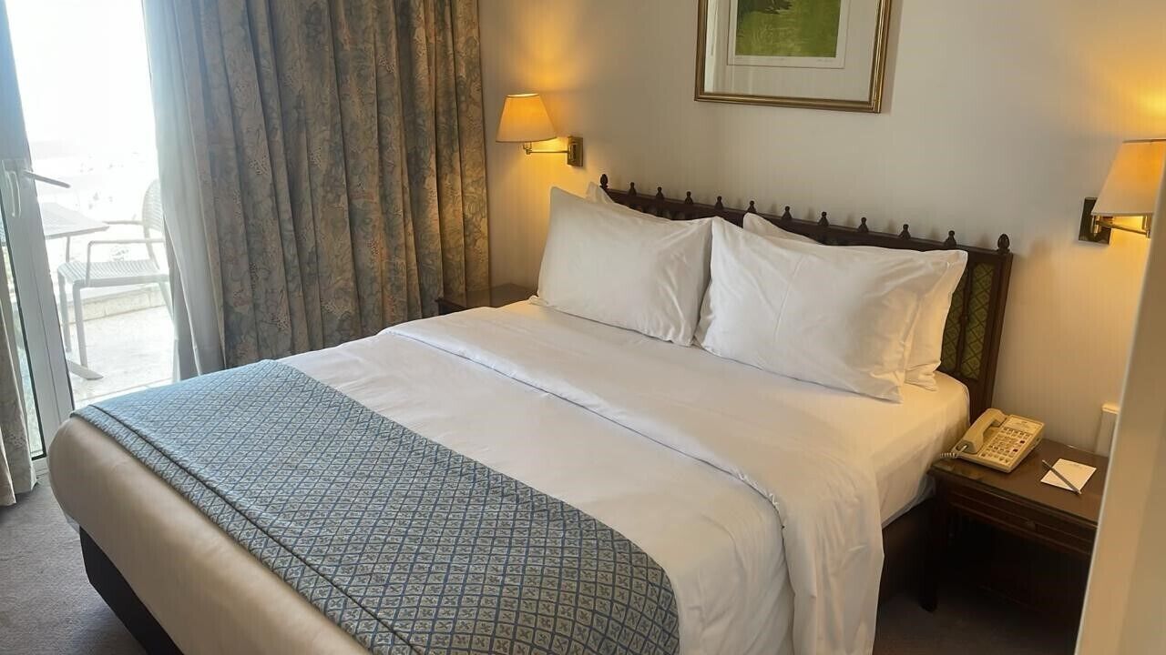 A hotel room with a bed, a bedside table with a phone, and a window with curtains.