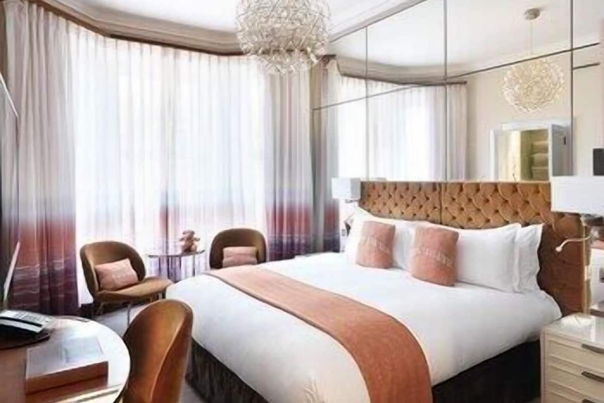A hotel room with a large bed, a round table, a chair, a chandelier, and a large window with curtains.