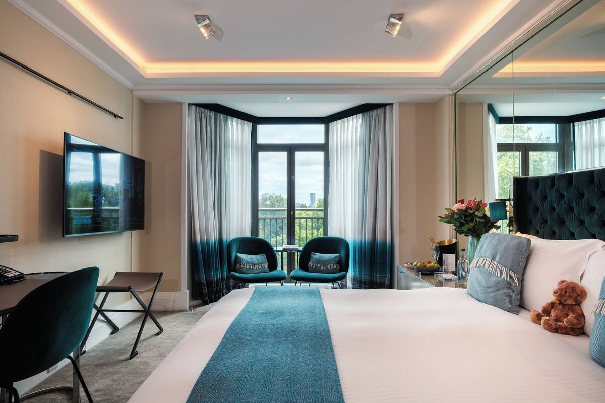 A hotel room with a large bed, a desk, two chairs, a television, and a view of the city through large windows.
