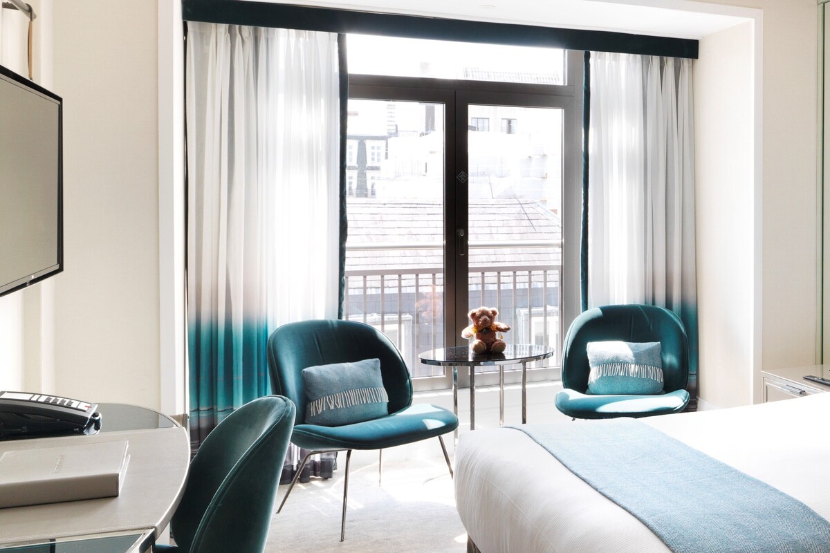 A hotel room with a bed, two teal chairs, a small table, a TV, and a large window with curtains.