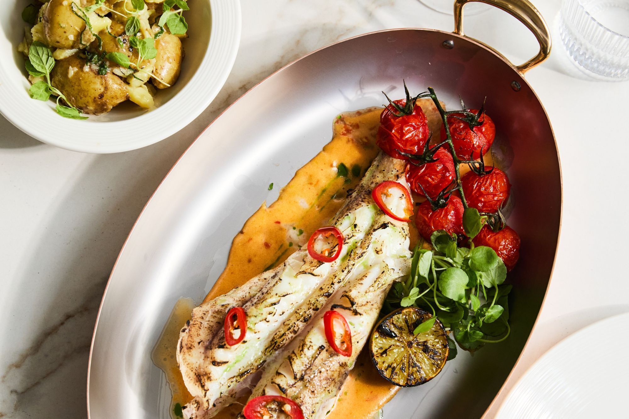 Un plato con pescado a la parrilla, tomates cherry y verduras de hoja verde.