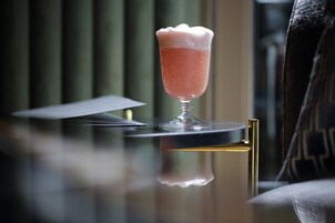 Ein Cocktailglas mit einem rosafarbenen Getränk auf einer spiegelnden Fläche, vermutlich einer Bar oder einem Tisch, mit unscharfem Hintergrund.