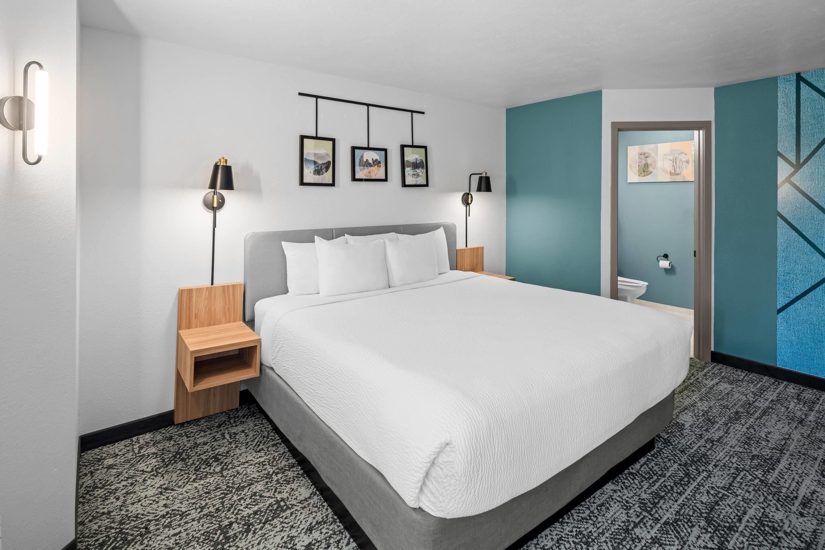 Habitación de hotel ordenada con una cama grande, mesitas de noche, una obra de arte en la pared y una puerta que da a otra habitación.