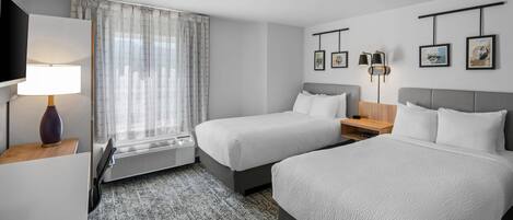 A hotel room with two beds, a desk, a TV, a heater, and framed pictures on the wall.