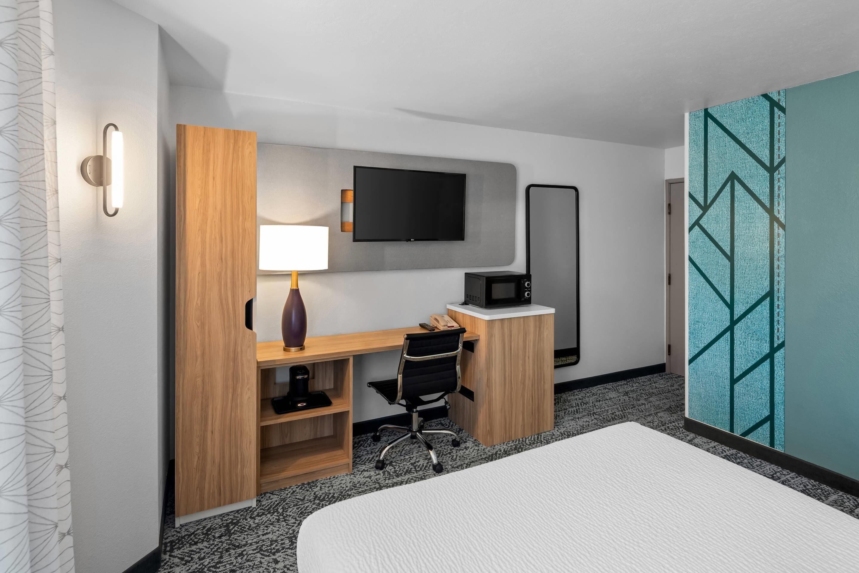 A hotel room with a bed, a desk with a chair, a TV, a microwave, and a large mirror.