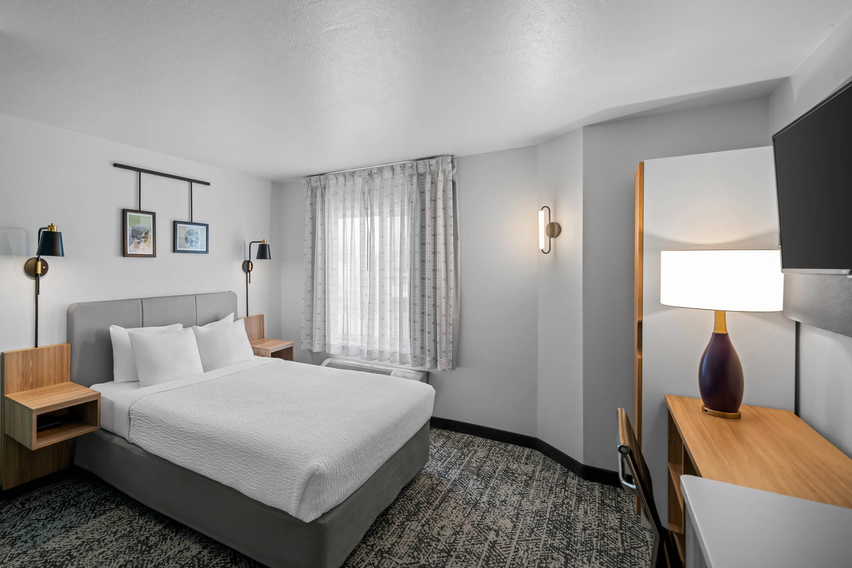 Une chambre d’hôtel comprenant un lit, des tables de chevet, un bureau, une chaise, une lampe, un téléviseur et une fenêtre avec des rideaux.